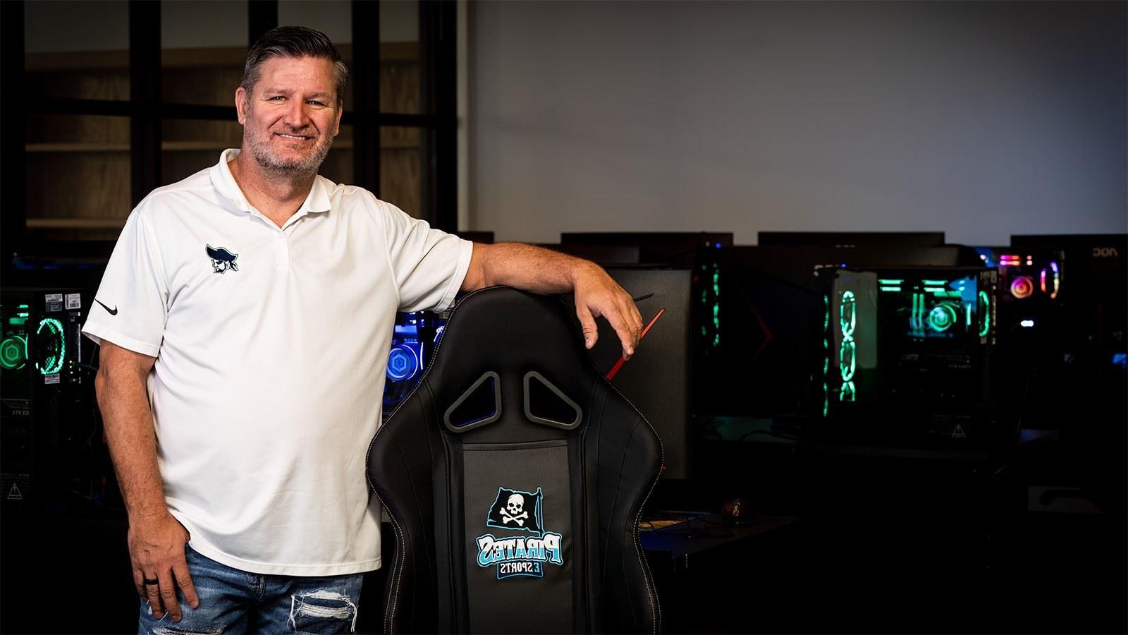 Scott Brumfield站在游戏电脑前. 他坐在PSC海盗队的游戏椅旁边，穿着海盗队的球衣.