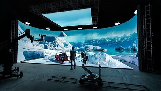 A man wearing jeans, sneakers, and a plaid shirt stands in a production studio. He is facing a large LED screen showing an icy mountain range and a snowmobile.