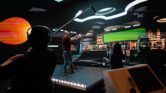 《十大菠菜台子》的导演杰森·墨菲站在一个虚拟宇宙飞船的舞台上. 他正在和一名工作人员交谈，另一名工作人员拿着吊杆麦克风.